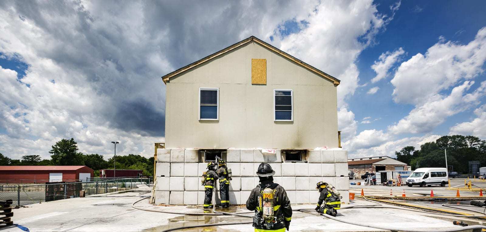 Firefighters in turnout gear flow a hoseline into a basement window of a two-story test house during fire experiments, while another firefighter watches from the yard.
