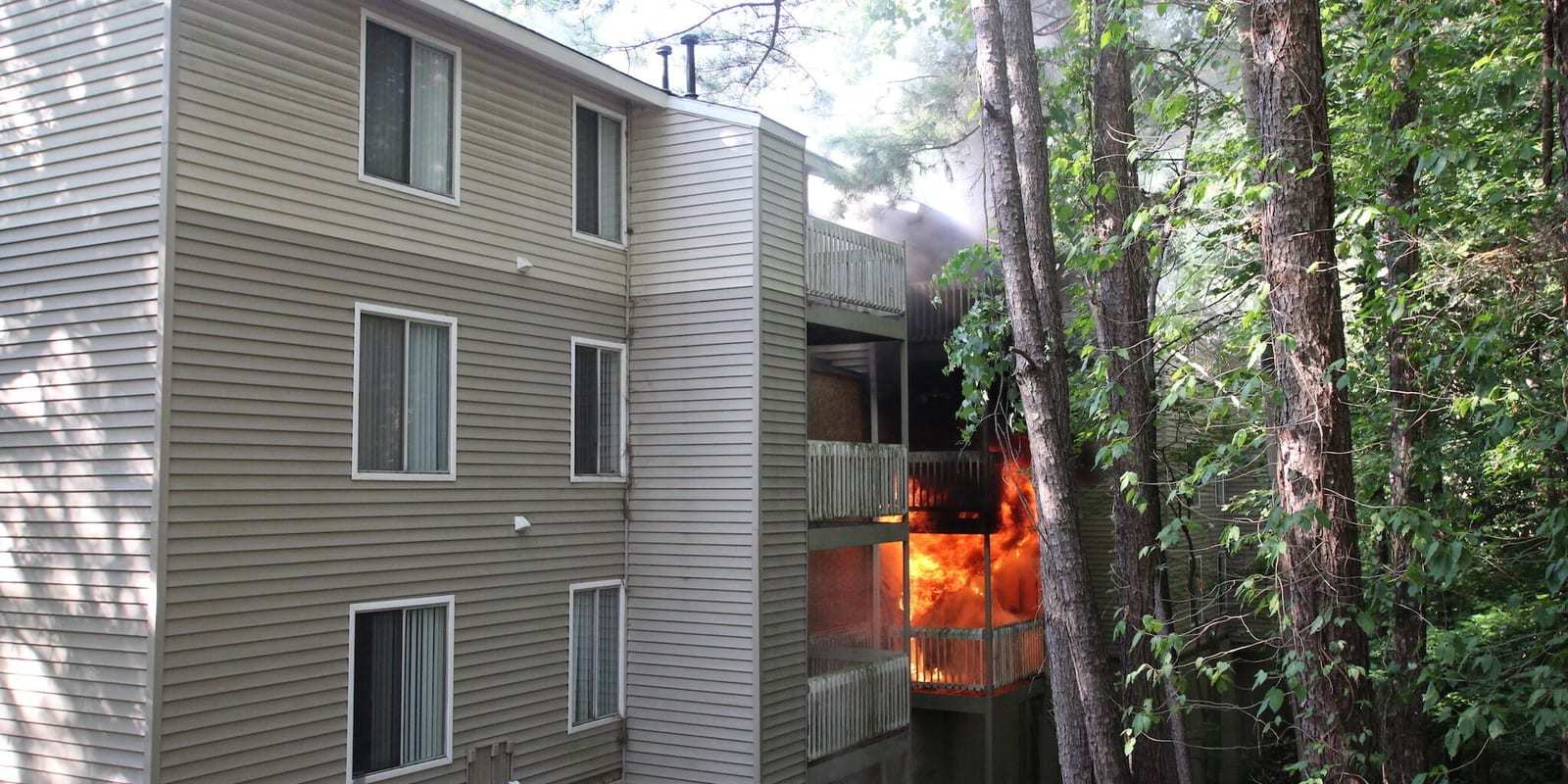 Fire burns on the balcony of a three-story garden-style apartment building with tan siding, surrounded by tall trees, during a research burn.