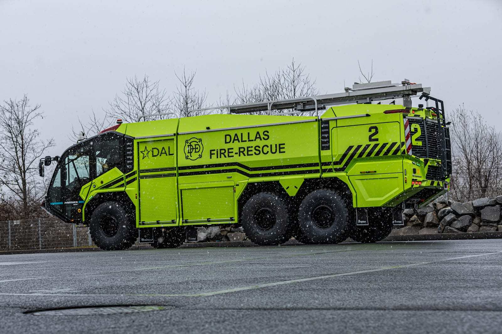 Lime-green Rosenbauer PANTHER 6x6 electric crash truck with DAL and Dallas Fire-Rescue markings, seen in a three-quarter side view on wet pavement with bare trees behind.