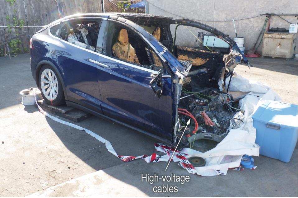 A blue SUV with its front end torn away and burned, and an arrow pointing to orange high-voltage cables exposed forward of the front seat.