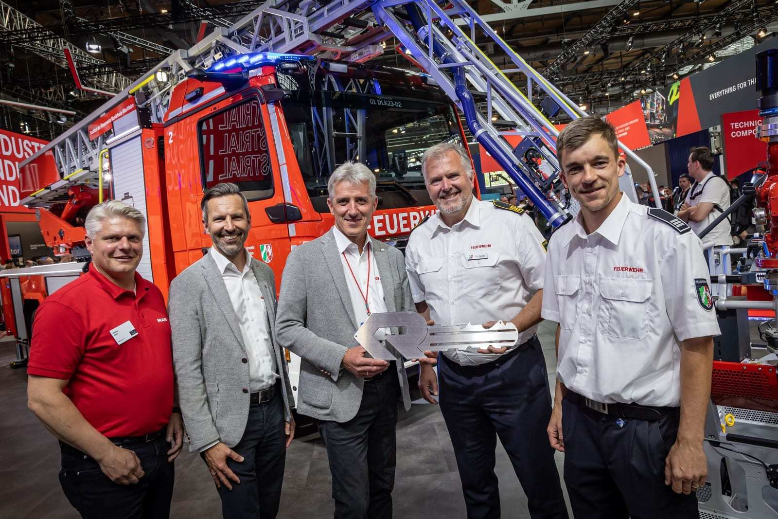 Rosenbauer, MAN and North Rhine-Westphalia fire institute representatives stand in front of the red L32A-XS electric aerial at the Interschutz 2026 trade show, one holding a metal handover plaque.