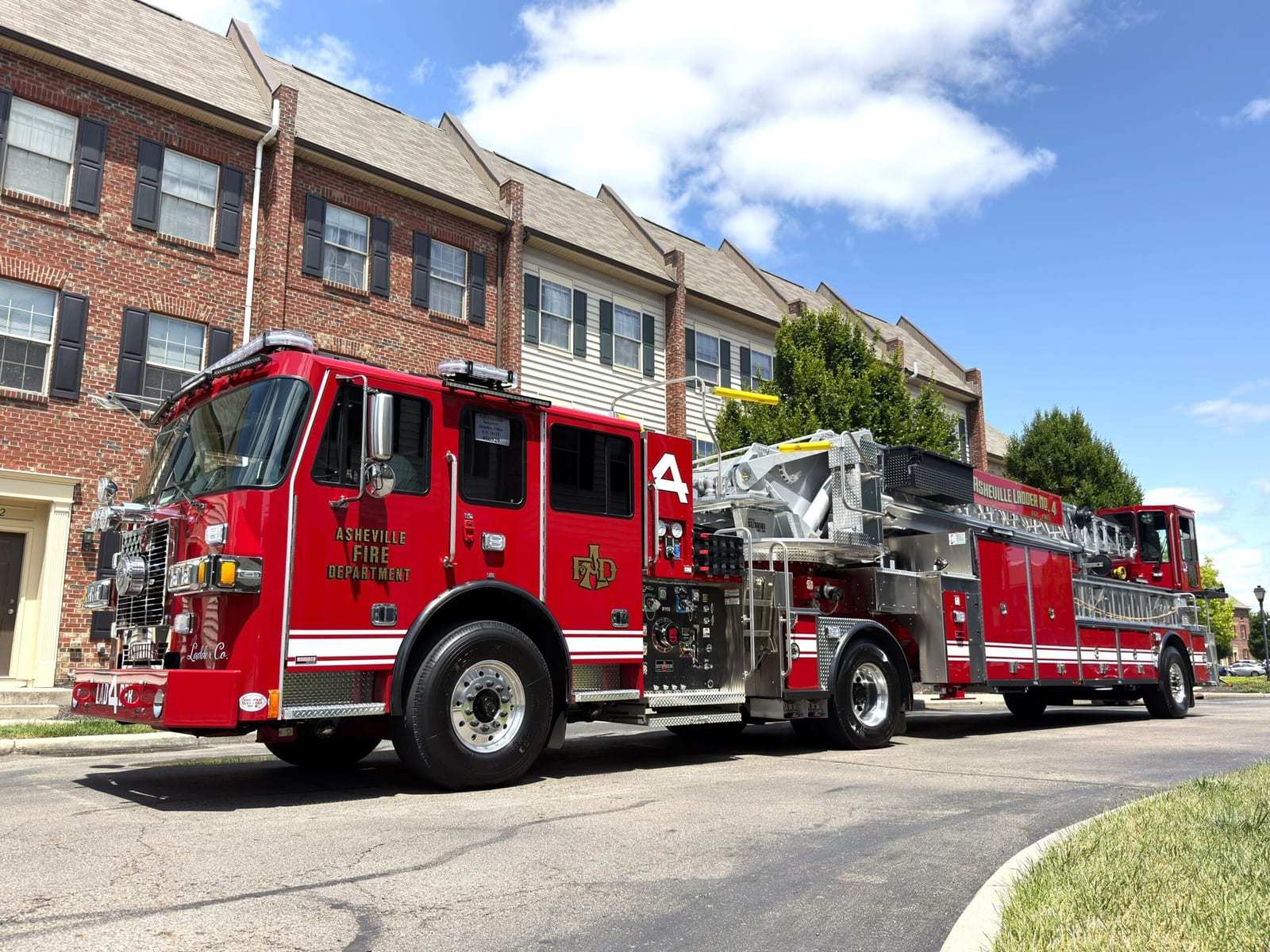 A red Asheville Fire Department tractor-drawn aerial with a stainless steel body and an enclosed tiller cab, parked on a residential street in front of brick townhouses.