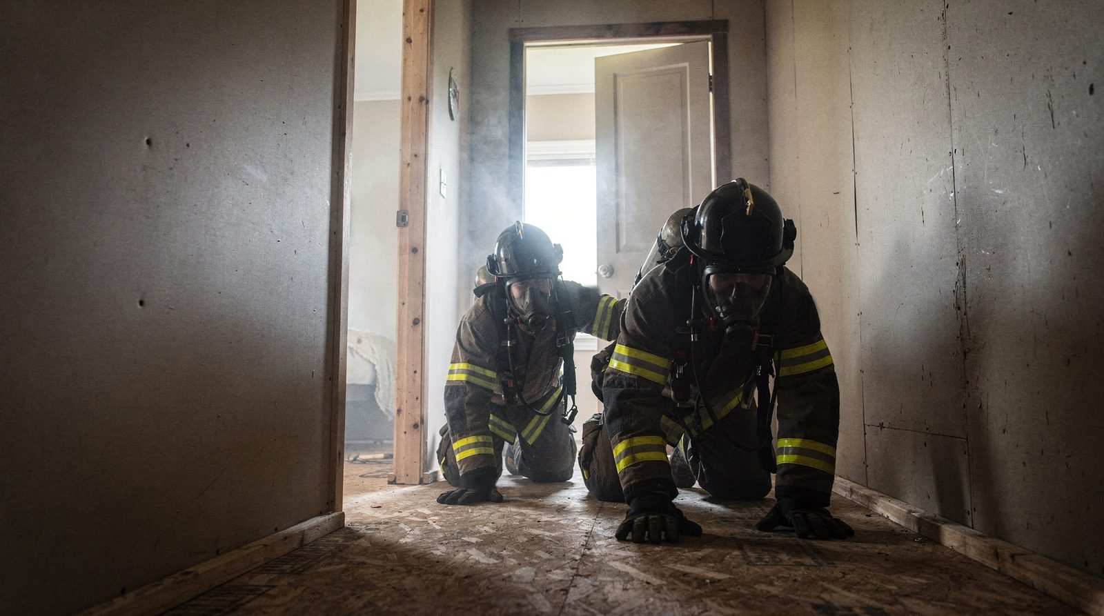Two firefighters in turnout gear and facepieces crawl a plywood hallway in a residential training prop, with daylight and light haze at an open door.