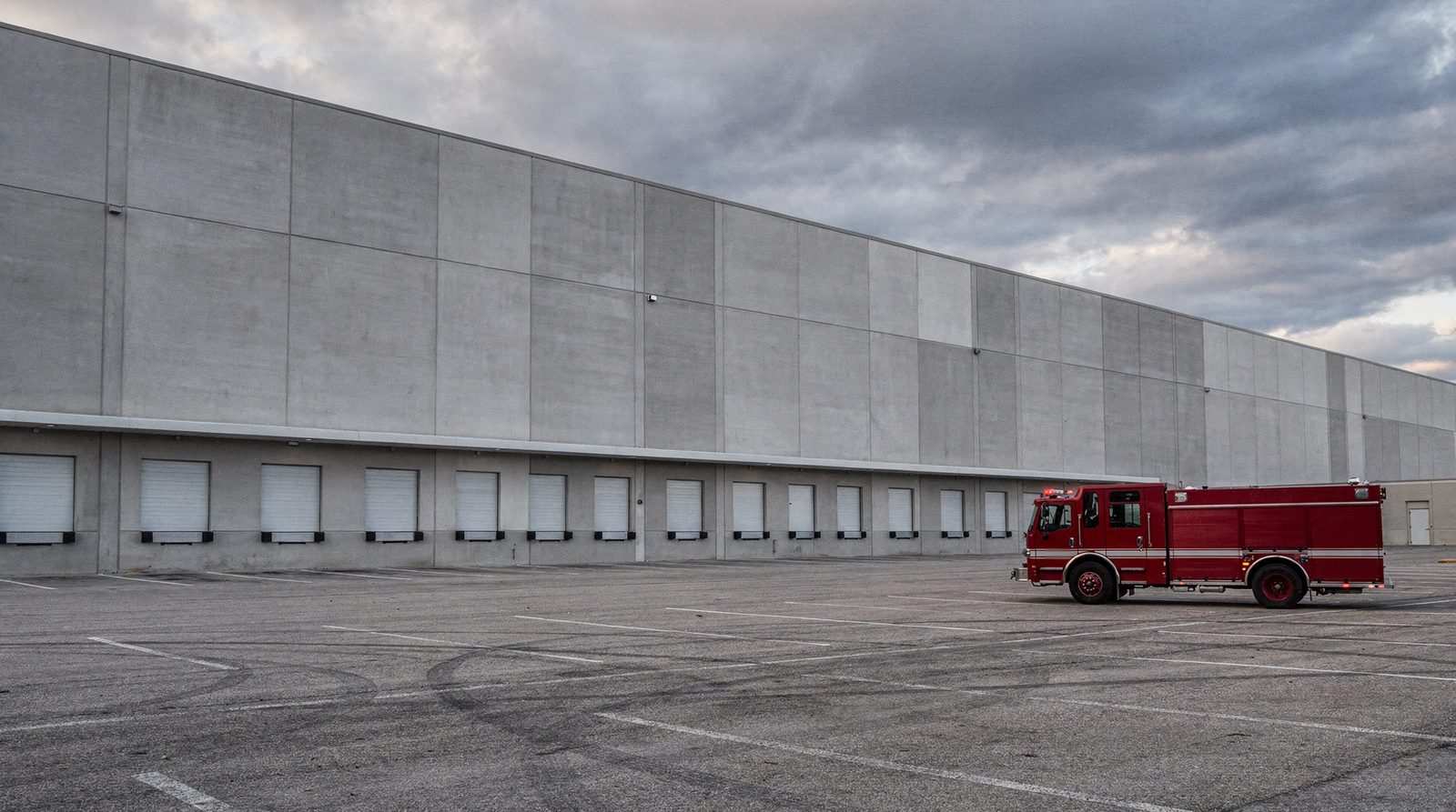 An unmarked fire engine with its lights off is parked in an empty lot in front of a long concrete warehouse with closed loading doors, under a cloudy daytime sky.